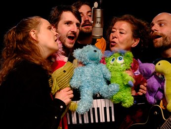 Fünf Personen singen in ein Mikrofon, umgeben von bunten Plüschtieren. Die fröhliche Gruppe wirkt in einem musikalischen Auftritt.