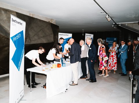 Eine Gruppe von Menschen steht an einem Tisch bei einer Veranstaltung. Banner im Hintergrund zeigen das Thema der Veranstaltung.