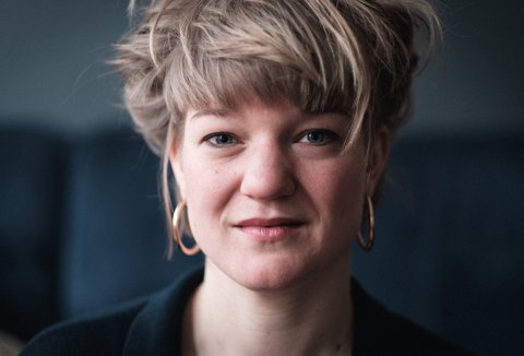 Porträt einer Person mit kurzen blonden Haaren und Ohrringen, die direkt in die Kamera schaut, vor einem unscharfen Hintergrund.