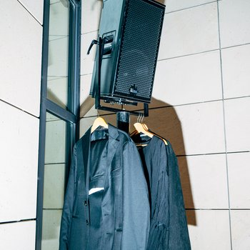 Ein Lautsprecher steht auf einem Ständer vor einer Wand. Zwei schwarze Sakkos hängen an ihm. Es gibt Schatten.