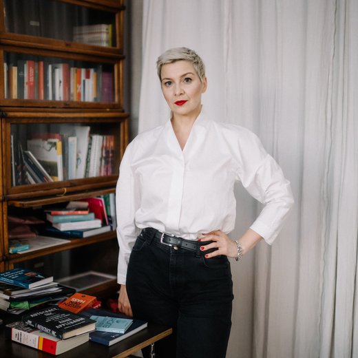 Eine Frau steht vor einem Bücherregal. Sie blickt in die Kamera. Ihre Haare sind kurz und sie trägt eine helle Bluse. Sie stützt den Arm in die Hüfte.
