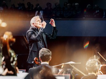 Ein Dirigent leitet ein Orchester, umgeben von sanftem Licht, während die Musiker konzentriert spielen.