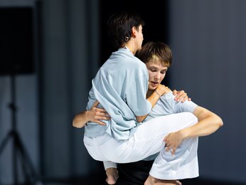 Zwei Tänzerinnen, eine hebt die andere in einer kraftvollen Pose auf einer minimalistisch beleuchteten Bühne. Beide sind barfuß.
