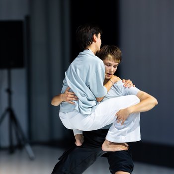 Zwei Tänzerinnen, eine hebt die andere in einer kraftvollen Pose auf einer minimalistisch beleuchteten Bühne. Beide sind barfuß.