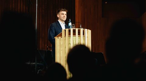 Ein Mann steht an einem Rednerpult vor einem Publikum. Der Raum ist holzgetäfelt und die Beleuchtung ist warm.