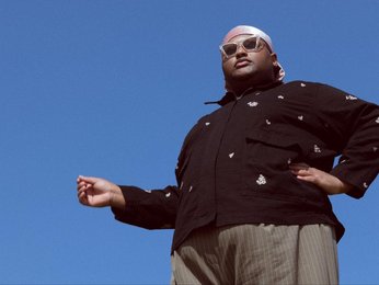 Ein Mann steht vor blauem Himmel, von unten fotografiert, mit einem dunklen Jacke, einem weißen Kopftuch und einer Sonnenbrille, eine Hand ausgestreckt.