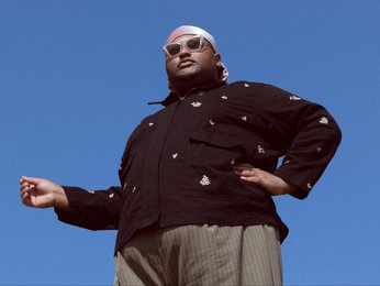Ein Mann steht vor blauem Himmel, von unten fotografiert, mit einem dunklen Jacke, einem weißen Kopftuch und einer Sonnenbrille, eine Hand ausgestreckt.