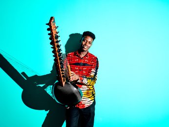 Ein Musiker in einem bunten Hemd spielt eine Kora vor einem hellblauen Hintergrund. Sein Gesichtsausdruck ist konzentriert.