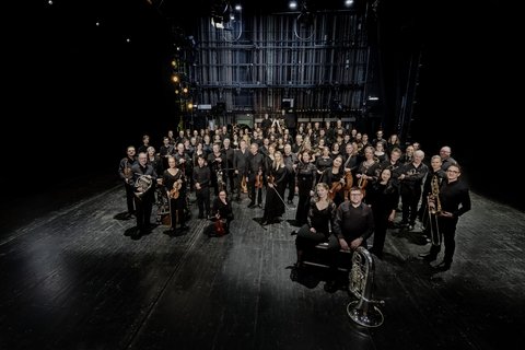 An orchestra is standing in a dark room, turned towards the camera. All are wearing concert clothes and holding instruments.