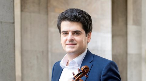 Ein Mann in einem blauen Anzug posiert draußen mit einer Violine in der Hand.