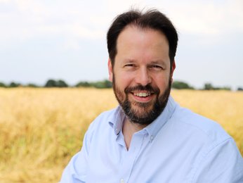 Ein Mann steht tagsüber vor einem Feld. Er trägt ein helles Hemd. Er blickt in die Kamera und lächelt.