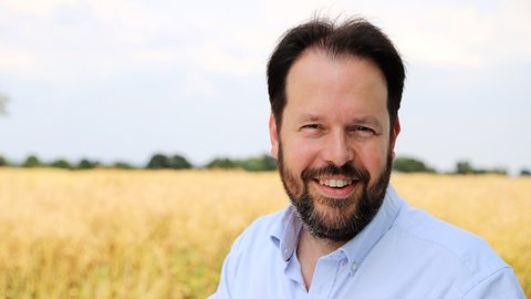 Ein Mann steht tagsüber vor einem Feld. Er trägt ein helles Hemd. Er blickt in die Kamera und lächelt. © Marshall Light Studio