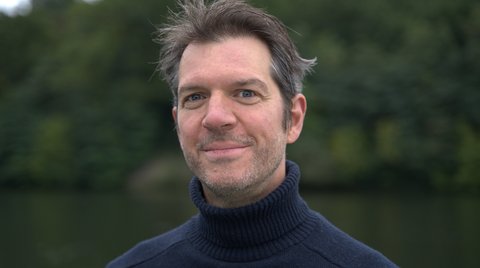 Ein Mann mit dunklem Rollkragenpullover lächelt vor einem Hintergrund aus Bäumen und einem Fluss.