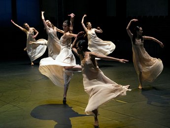 Eine Gruppe von Tänzerinnen in fließenden Kleidern führt eine elegante Choreografie auf einer schattigen Bühne auf.