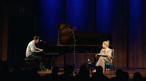 Ein Pianist spielt auf einer Bühne mit einem Flügel, während eine Person auf einem Stuhl sitzt und zuhört.