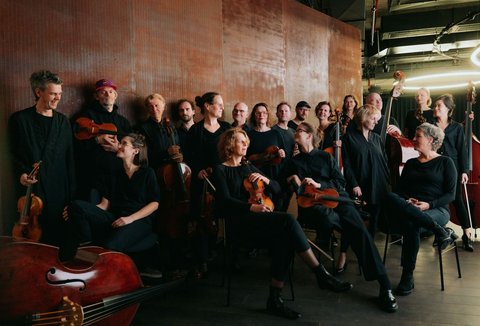 A group of people is standing standing and sitting in varying positions. Everybody is holding an instrument. They are looking in different directions and smiling or talking.