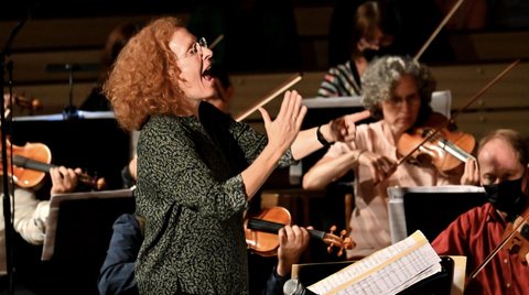 Eine Dirigentin mit lockigem Haar leitet ein Orchester. Mehrere Musiker spielen Streicherinstrumente, konzentriert auf die Musik.