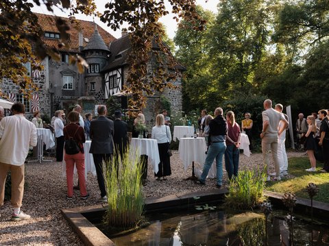 Menschen stehen draußen in einem eleganten Garten bei einem gesellschaftlichen Event. Ein historisches Gebäude ist im Hintergrund sichtbar.