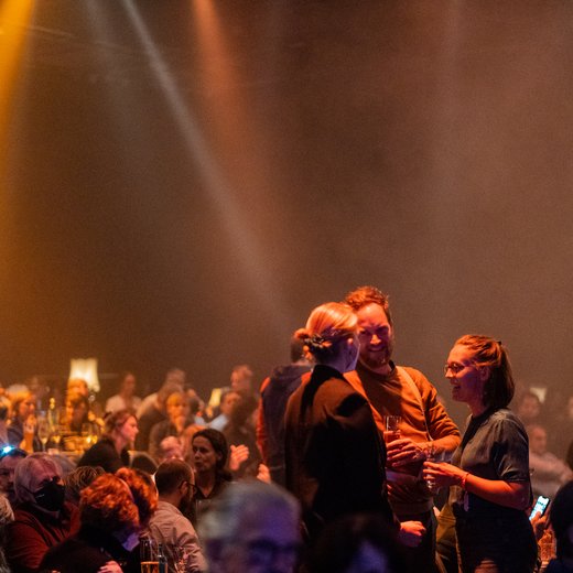 Menschen auf einer gut besuchten Veranstaltung in einem dunklen Saal mit warmem Licht. Sie unterhalten sich und genießen die Stimmung.