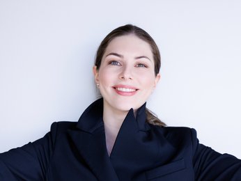 Eine Frau in einem dunklen Anzug lächelt mit ausgestreckten Armen. Sie steht vor einem hellen Hintergrund.