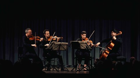 Ein Streichquartett spielt auf einer Bühne unter gedämpftem Licht. Vier Musiker mit Notenblättern sind auf klassische Instrumente fokussiert.