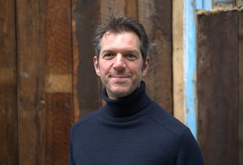 A man in a dark blue turtleneck stands in front of a wooden wall. He is smiling softly, with dark hair.