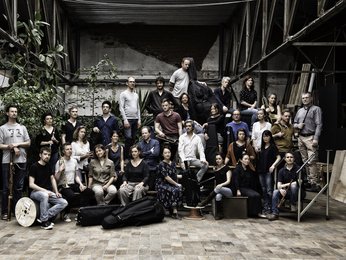 Eine große Gruppe von Menschen posiert gemeinsam in einem industriellen Raum. Einige halten Musikinstrumente, andere stehen lässig.